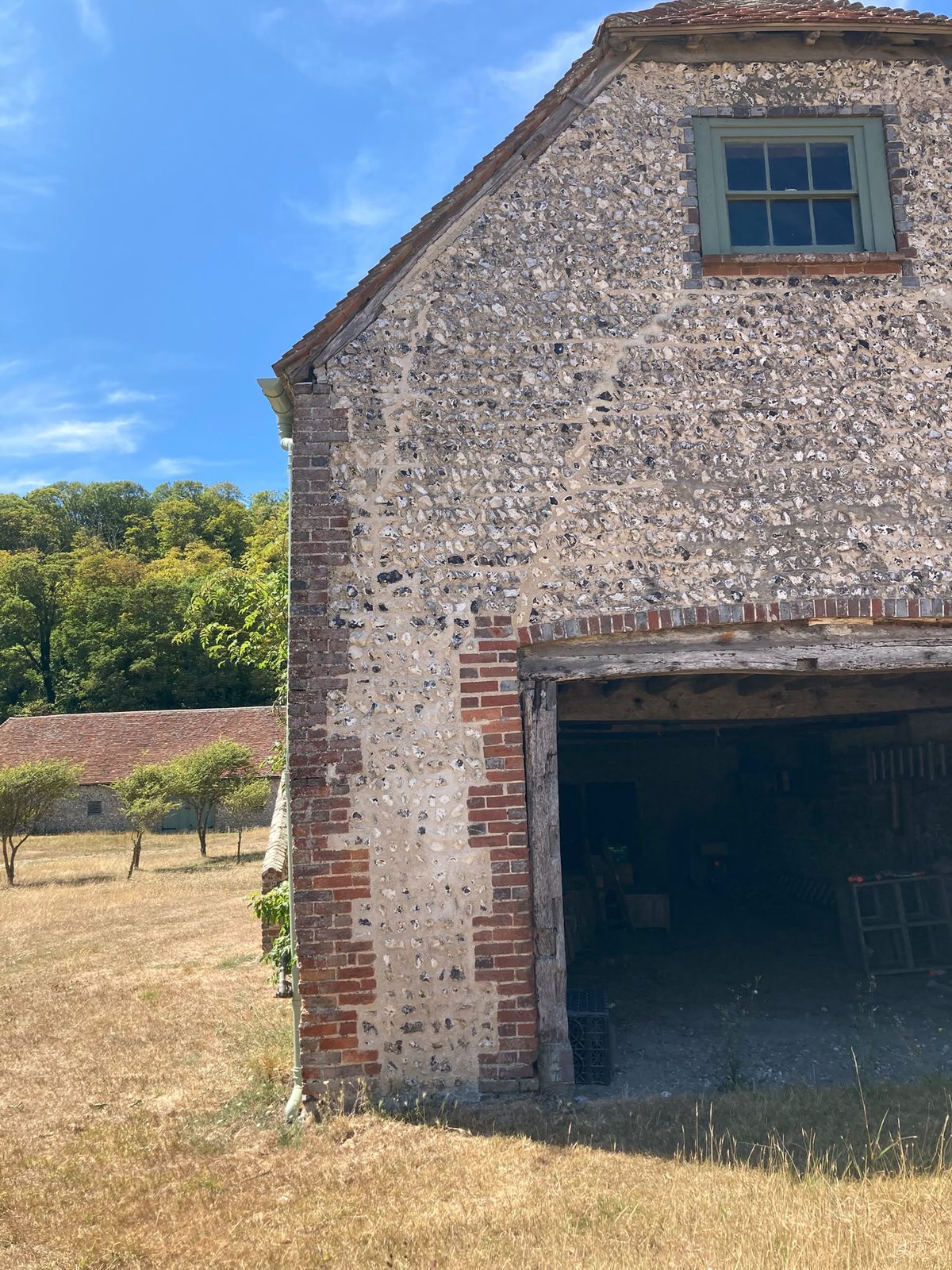 A flint barn with brick quoins, a green casement window and an open doorway, standing in a dry summer field under a blue sky