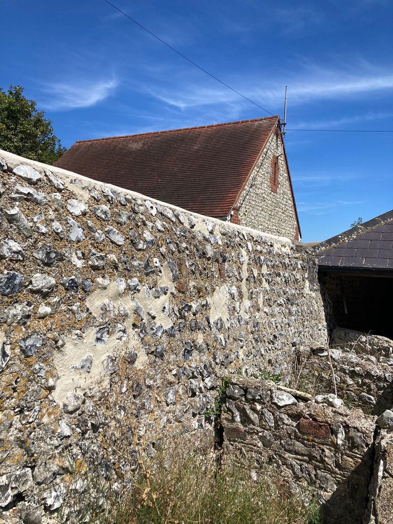 A flint wall with a tiled roof and gable beyond, against a blue sky